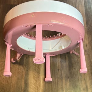 Op de afbeelding: Een roze en witte ronde breimachine met vier poten. De poten zijn gemaakt van roze plastic en op een van de poten staat "NWCC" gedrukt.