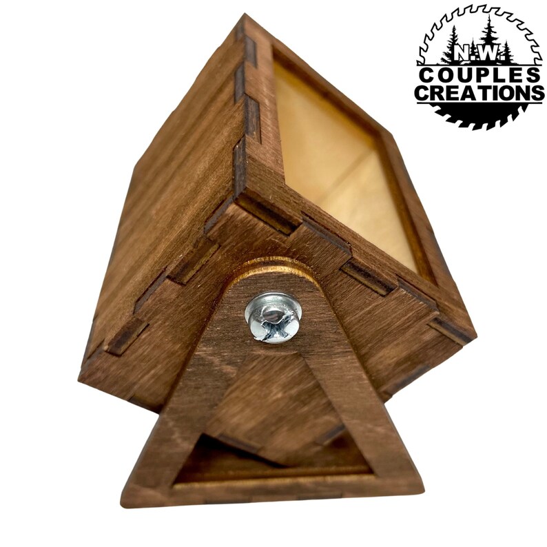 May include: A wooden box with a hinged lid and a small metal screw. The box is stained a dark brown color and has a light wood interior. The box is sitting on a triangular base.