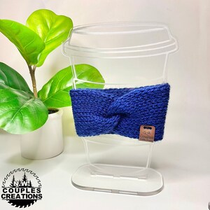 May include: Blue knitted coffee cup cozy with a brown tag that says "A Couples Stitches". The cozy is displayed on a clear acrylic coffee cup holder.