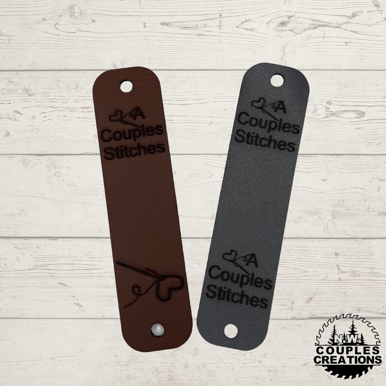May include: Two leather bookmarks with a heart and the text "A Couples Stitches" on them. The bookmarks are brown and gray. The bottom bookmark has a small needle and thread design.