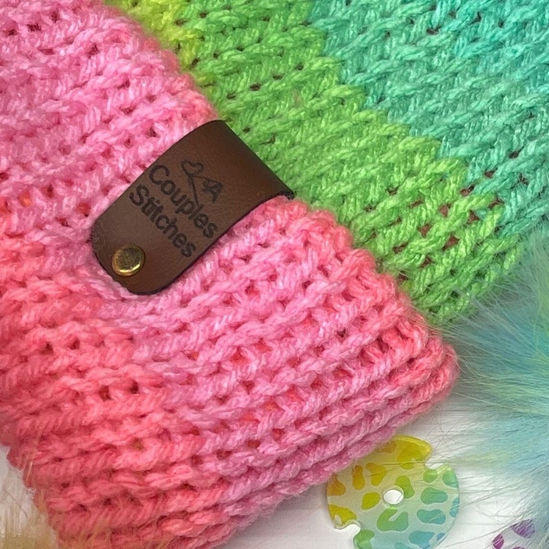 May include: A pink, green, and yellow striped crocheted headband with a brown leather tag that says "Couples Stitches".