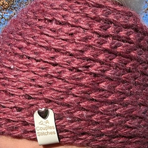 May include: A burgundy knitted hat with a white pom pom on top. The hat has a small white leather tag attached to it with a silver heart and the text "2 A Couples Stitches".