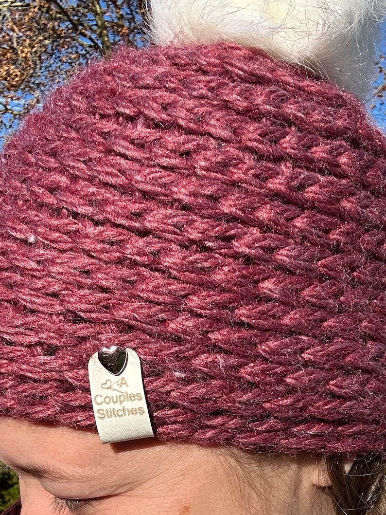 May include: A burgundy knitted hat with a white pom pom on top. The hat has a small white leather tag attached to it with a silver heart and the text "2 A Couples Stitches".