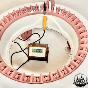以下が含まれることがあります： ピンクと白の円形の編み機をクローズアップした写真。木製のタイマーが「00006」を表示しており、タイマーは黄色のワイヤーに接続されています。