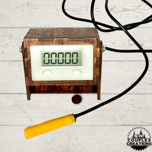 May include: A wooden box with a digital counter displaying "00000". The box has two white buttons and a black wire attached to it. A yellow probe is lying on a white wooden surface.