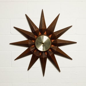 May include: A brown wooden starburst clock with a gold face and black hands. The clock has a 12-hour face with Roman numeral markings.