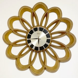 May include: A gold-toned wall clock with a flower-shaped frame and black Roman numeral clock face. The clock face has a silver background and black hands.
