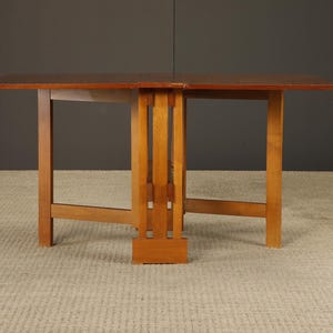 May include: A wooden drop-leaf table with a rectangular top and a light brown finish. The table has a sturdy base with vertical supports and a central support structure. The table is designed to fold down for storage.
