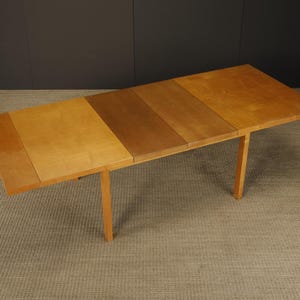May include: A wooden rectangular table with a light brown finish. The table has a multi-panel top with varying shades of brown and is supported by four straight legs. The table is approximately 60 inches long.