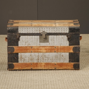 May include: A rectangular storage trunk featuring a light gray patterned metal body with horizontal wooden slats and dark metal corner reinforcements. The front includes a central metal latch mechanism and a keyhole plate with dark metal trim.