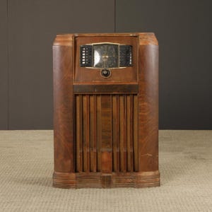 May include: An antique wooden radio cabinet with a dark brown finish. The radio features a dial with a gold-colored frame, black speaker vents, and vertical wooden slats. The cabinet has a rectangular shape with rounded edges.