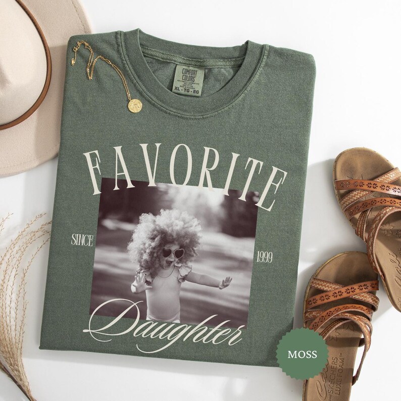May include: A moss green t-shirt with the words "FAVORITE DAUGHTER" printed on the front, along with a black and white photo of a child. A gold necklace and brown sandals are also visible.