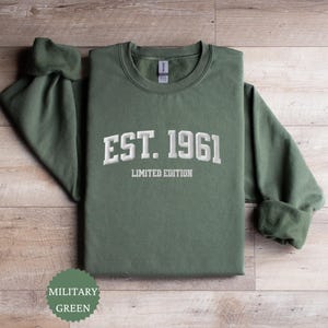 May include: A military green sweatshirt with the text "EST. 1961 LIMITED EDITION" in white. The sweatshirt is displayed on a wooden surface. The sleeves are folded at the cuffs. A circular label at the bottom left corner reads "MILITARY GREEN".