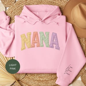 May include: Pale pink hoodie featuring the word "NANA" embroidered in pastel colours. The sleeve displays the names "Liam, Olivia, Diana" in a script font. The hoodie is presented on a woven mat, next to a brown hat.