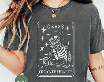 Puede incluir: Camiseta gris oscuro con un gráfico blanco de un esqueleto sosteniendo una taza, dentro de un marco rectangular con estrellas y fases lunares. El texto "THE OVERTHINKER" está debajo. La camiseta es color pimienta.