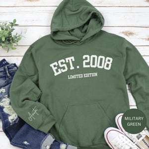 May include: Military green hoodie with the text "EST. 2008 LIMITED EDITION" in white. The hoodie has a front pocket and a drawstring hood. A pair of blue jeans and white sneakers are next to the hoodie.