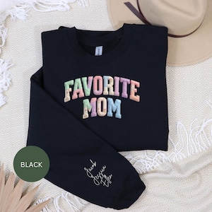Può includere: Felpa nera a maniche lunghe con le parole "FAVORITE MOM" in colori pastello. La manica ha i nomi "Jacob, Jessica, Ellen" in corsivo. Un cerchio nero con la parola "BLACK" è nell'angolo in basso a sinistra. Un cappello beige è sullo sfondo.