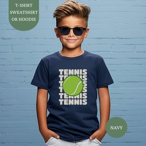 Può includere: Una maglietta blu navy con la parola "TENNIS" ripetuta cinque volte e un'immagine di una palla da tennis verde. La maglietta è indossata da un bambino che indossa occhiali da sole. Il testo "NAVY" è visibile nell'angolo in basso a destra dell'immagine.