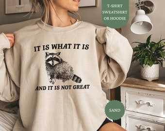 May include: A beige sweatshirt with the text "IT IS WHAT IT IS AND IT IS NOT GREAT" and a black and white illustration of a raccoon. The sweatshirt is available in a sand color.