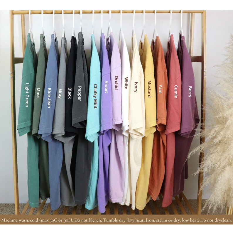 May include: A wooden clothing rack displays a variety of solid-colored t-shirts. Colors include light green, moss, blue jean, gray, black, pepper, chalky mint, violet, orchid, white, ivory, mustard, yam, cumin, and berry. Each shirt has a label with its color name.