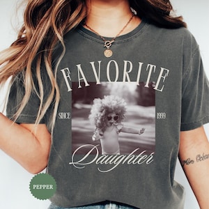 May include: Dark gray t-shirt with the words "FAVORITE Daughter" printed on the front, along with a black and white photo of a child. The text "SINCE 1999" is also printed on the shirt. The shirt is a Pepper color.