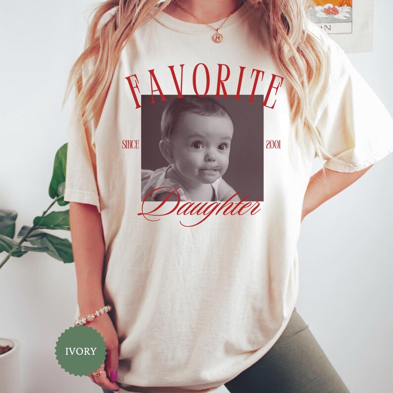 May include: Ivory-colored t-shirt featuring a black and white photo of a baby with the words "FAVORITE Daughter" in red script. The text "SINCE 2001" is also visible. The shirt has a relaxed fit.