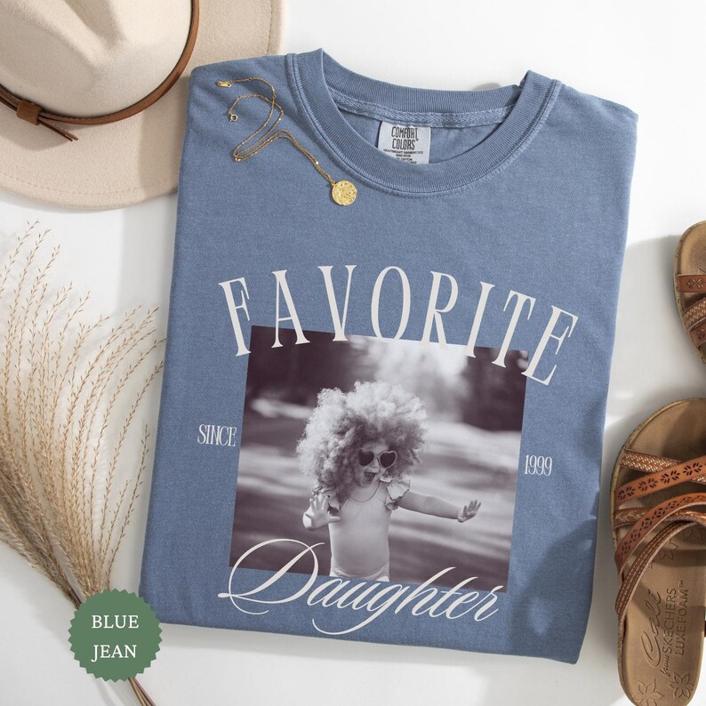 May include: Blue jean Comfort Colors t-shirt with the words "FAVORITE Daughter" and a black and white photo of a child. A gold necklace and a beige hat are next to the shirt. The shirt has the text "SINCE 1999".