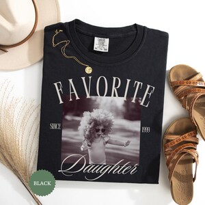 May include: Black crew neck sweatshirt with the words "FAVORITE DAUGHTER" printed in white. The sweatshirt features a black and white photo of a child wearing sunglasses. A gold necklace and brown sandals are also in the image.