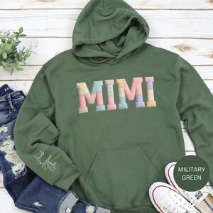 Könnte beinhalten: Militärgrüner Kapuzenpullover mit dem Wort "MIMI" in pastellfarbenen, gesteppten Buchstaben auf der Brust. Der Hoodie hat eine Kapuze mit Kordelzug, eine Vordertasche und den Text "Jacob Jessica Ella" auf dem linken Ärmel.