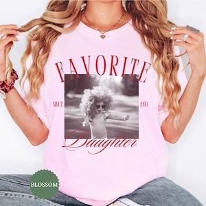 May include: A light pink t-shirt with the words "FAVORITE Daughter" in red script. A black and white photo of a child wearing sunglasses is in the center. The text "SINCE 1999" is also on the shirt. A green logo is at the bottom.