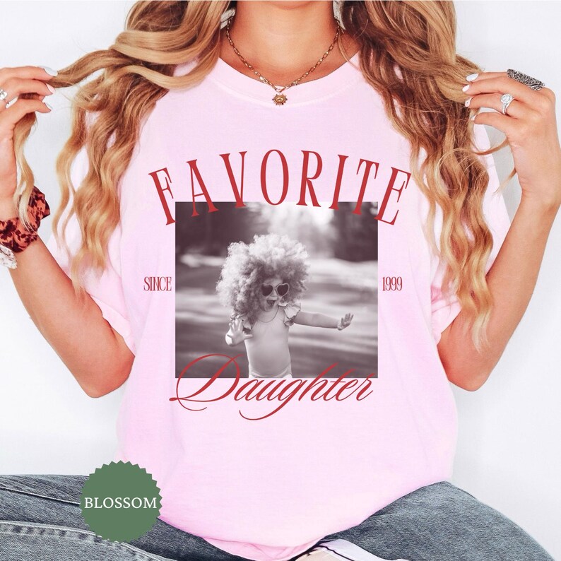 May include: A light pink t-shirt with the words "FAVORITE Daughter" in red script. A black and white photo of a child wearing sunglasses is in the center. The text "SINCE 1999" is also on the shirt. A green logo is at the bottom.