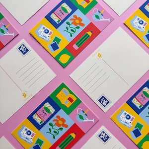 Könnte beinhalten: Eine Sammlung farbenfroher Postkarten mit verschiedenen Illustrationen, darunter eine Kamera, eine Pastadose, eine Blume, ein Bleistift und eine Zitrone. Die Postkarten sind auf einer rosa Oberfläche angeordnet, mit einer weißen Rückseite und Linien zum Schreiben.