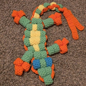 May include: A colourful, handmade crochet lizard toy. The lizard has a green body with yellow and blue details, and orange legs and tail. The toy is made with a textured stitch and is on a grey carpet.