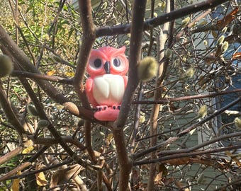 May include: A pink owl figurine with white accents and black eyes perched on a branch of a bare tree with buds.