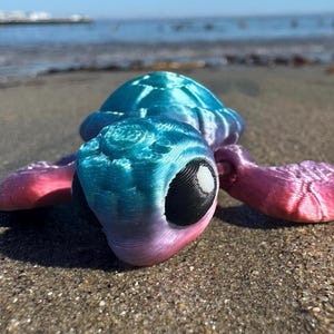 May include: A small, 3D printed sea turtle toy with a blue, green, and pink gradient color scheme. The turtle has a large, black eye and a white highlight. The toy is lying on a sandy beach.
