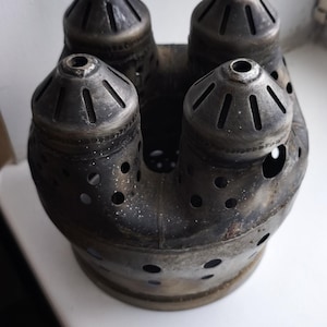 Peut inclure: Objet métallique sombre et vieilli, avec quatre becs coniques et une chambre centrale. Il présente de nombreux petits trous et une apparence usée. Posé sur une surface blanche, il semble ancien.