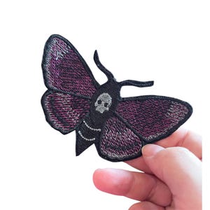 May include: An embroidered moth patch featuring a black body with a white skull design on the thorax. The wings are primarily dark purple with black edges and intricate black stitching details throughout the wing surface, creating a textured appearance.