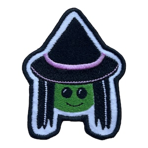 May include: Embroidered patch with a cartoon witch design. The witch has a green face, large eyes, and a smiling mouth. She wears a black witch hat with a pink band and black hair. The patch is outlined in white.