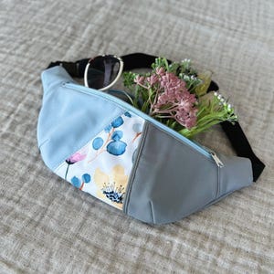 May include: A light blue and gray fanny pack with a floral design. The fanny pack has a black strap and a silver zipper. Sunglasses and flowers are inside the fanny pack.