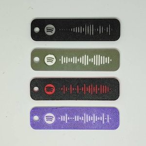 Könnte beinhalten: Vier verschiedene farbige Schlüsselanhänger mit einem Spotify-Logo und einem Barcode. Die Schlüsselanhänger sind schwarz, grün, schwarz mit rotem Logo und lila.