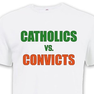 Puede incluir: Camiseta blanca con el texto "CATHOLICS" en verde sobre "VS." y "CONVICTS" en naranja. La camiseta tiene mangas cortas y cuello redondo.