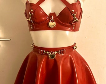 May include: A red latex skirt with a gold chain belt and a heart-shaped padlock. The skirt is a-line and has a ruffled hem. The top is a red latex bra with gold hardware and a heart-shaped padlock.