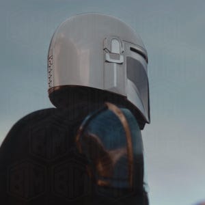 May include: A silver Mandalorian helmet with a dark visor and a distinctive T-shaped opening. The helmet is part of a suit of armor, with blue and gold accents visible on the shoulder. The image is a close-up, focusing on the helmet's details.