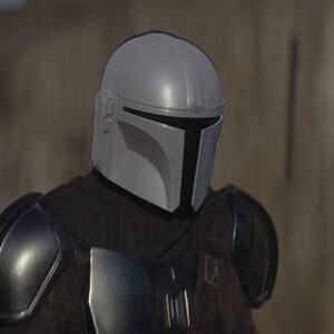 May include: A silver-colored Mandalorian helmet with a T-shaped visor. The helmet is part of a costume, with a dark gray outfit and shoulder armor visible. The helmet has a smooth, metallic finish.