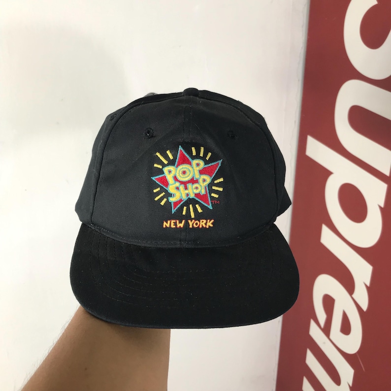 May include: Black baseball cap with a flat brim. The cap features an embroidered design of a star with the words "POP SHOP NEW YORK" in colorful lettering. The cap is made of a durable fabric.