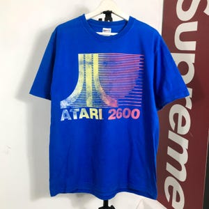 Puede incluir: Una camiseta azul brillante de manga corta con un estampado gráfico del logotipo de Atari y el texto ATARI 2600. El diseño gráfico consiste en un patrón degradado estilizado en amarillo, naranja y rosa con líneas horizontales hacia la derecha.
