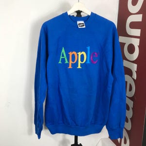 以下が含まれることがあります： 胸元にマルチカラーのレインボーフォントでAppleという文字が刺繍された、ロイヤルブルーの長袖スウェットシャツ。この衣類はクルーネックで、袖口と裾はリブ編みになっており、襟元には白いブランドタグが見えます。