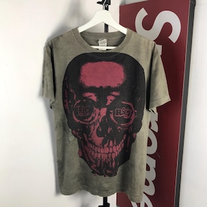 May include: Olive green t-shirt featuring a large skull graphic in black and burgundy. The skull design includes the letters "RSC" in the eye sockets. The t-shirt is hanging on a white hanger.