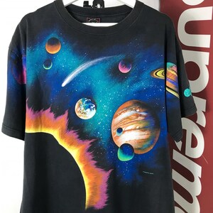 Puede incluir: Camiseta negra con un gr&aacute;fico colorido con tem&aacute;tica espacial. El dise&ntilde;o incluye planetas, un cometa y un sol brillante sobre un fondo azul oscuro y verde azulado. La camiseta tiene cuello redondo y mangas cortas.