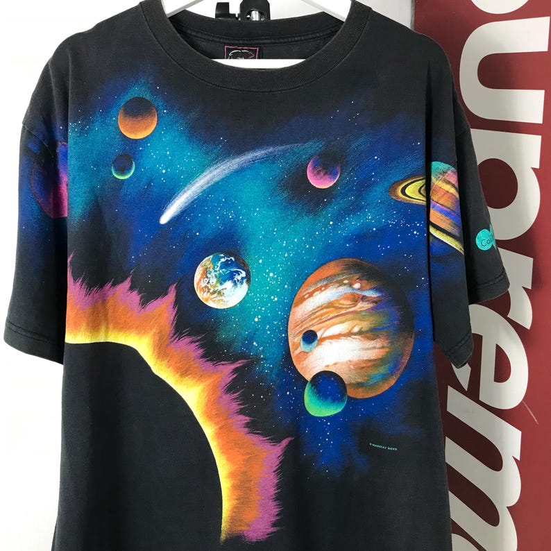 Puede incluir: Camiseta negra con un gr&aacute;fico colorido con tem&aacute;tica espacial. El dise&ntilde;o incluye planetas, un cometa y un sol brillante sobre un fondo azul oscuro y verde azulado. La camiseta tiene cuello redondo y mangas cortas.
